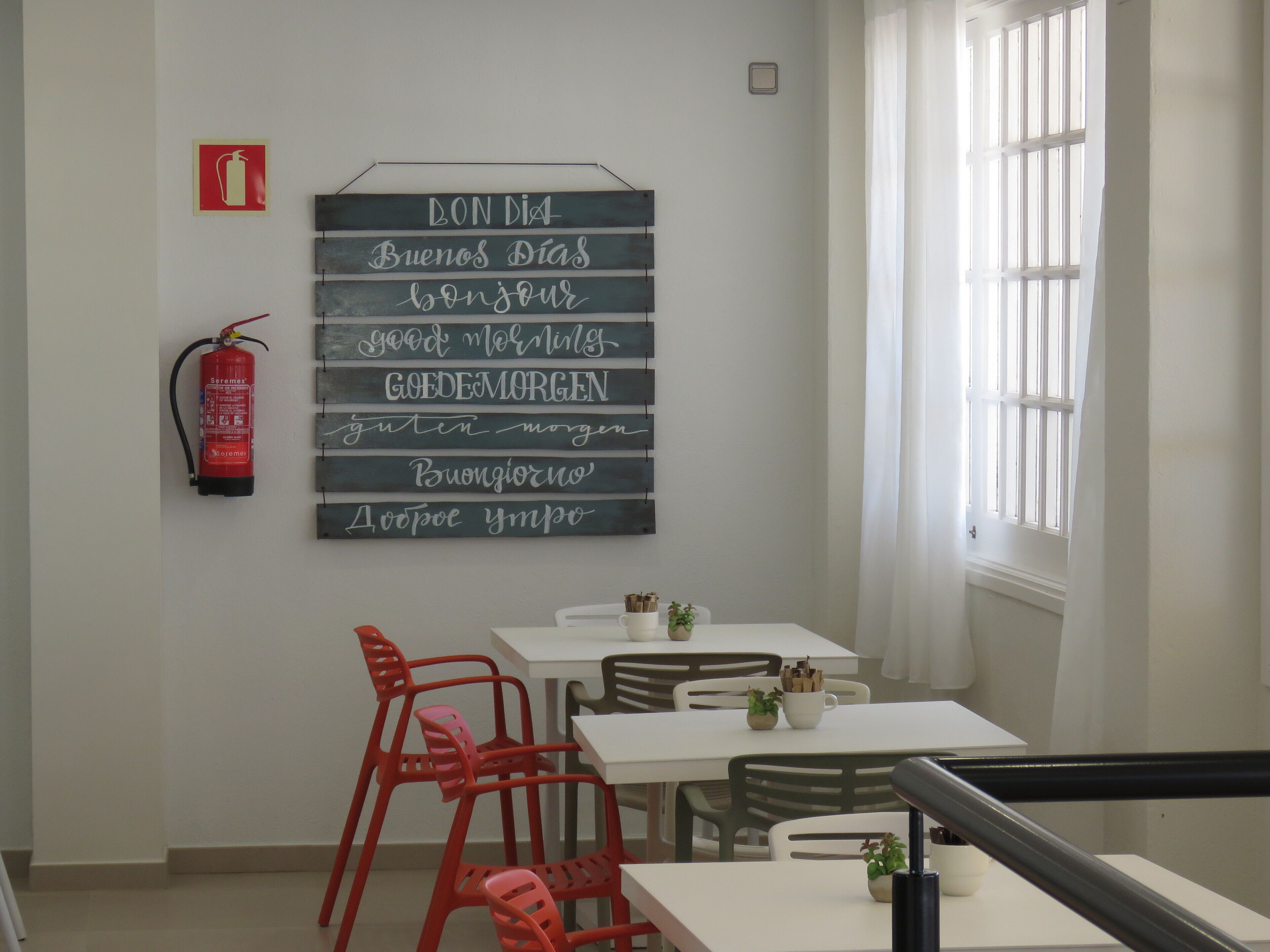 Una zona de menjador amb un extintor, un senyal amb salutacions en diversos idiomes i una finestra amb persianes.