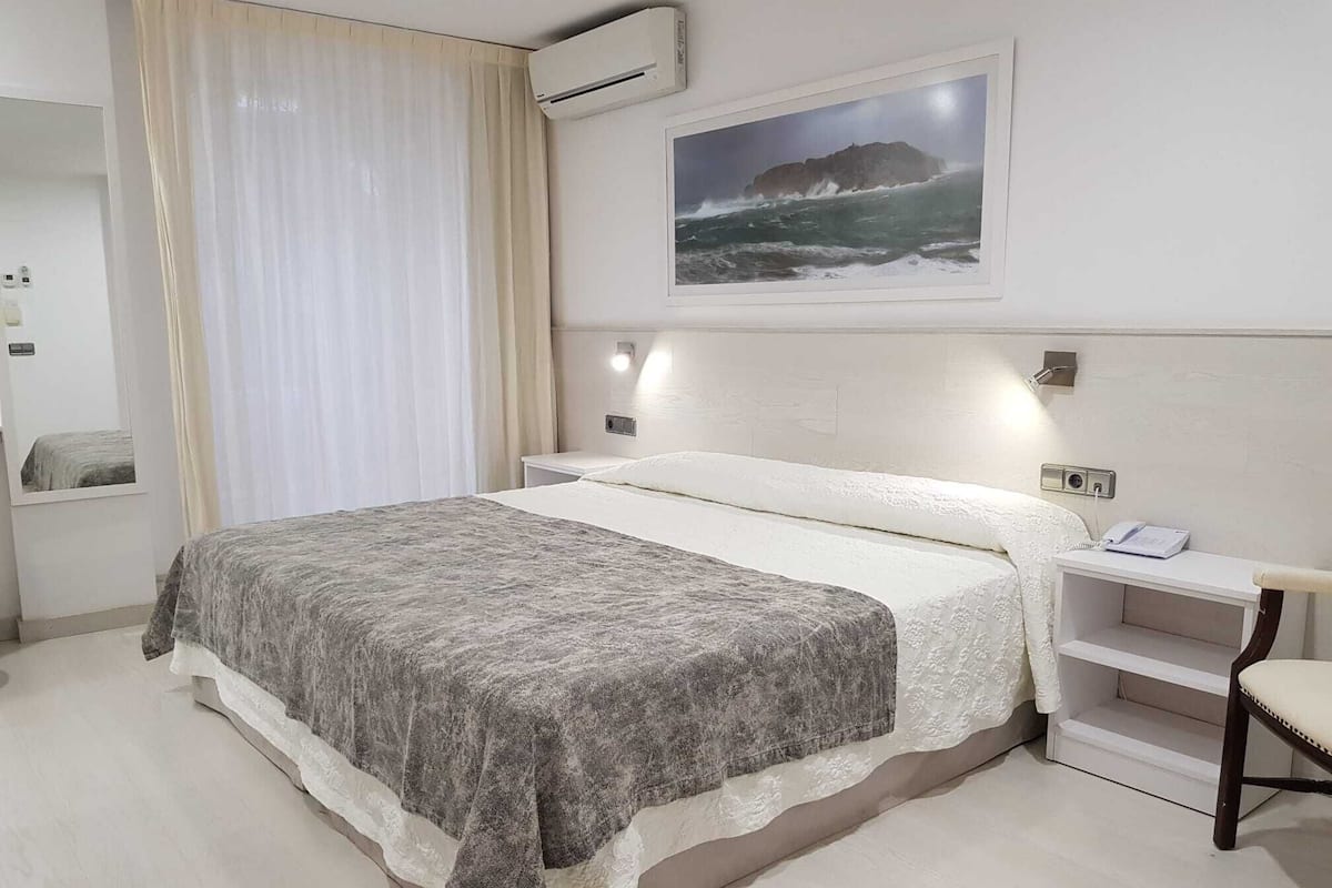 A hotel room with a bed, a bedside table with a phone, a picture on the wall, and an air conditioning unit.