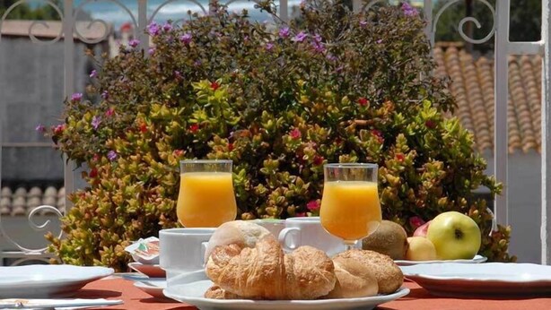 Ein Frühstückstisch mit Croissants, Äpfeln und Orangensaft, mit Blick auf das Meer und einen Garten.