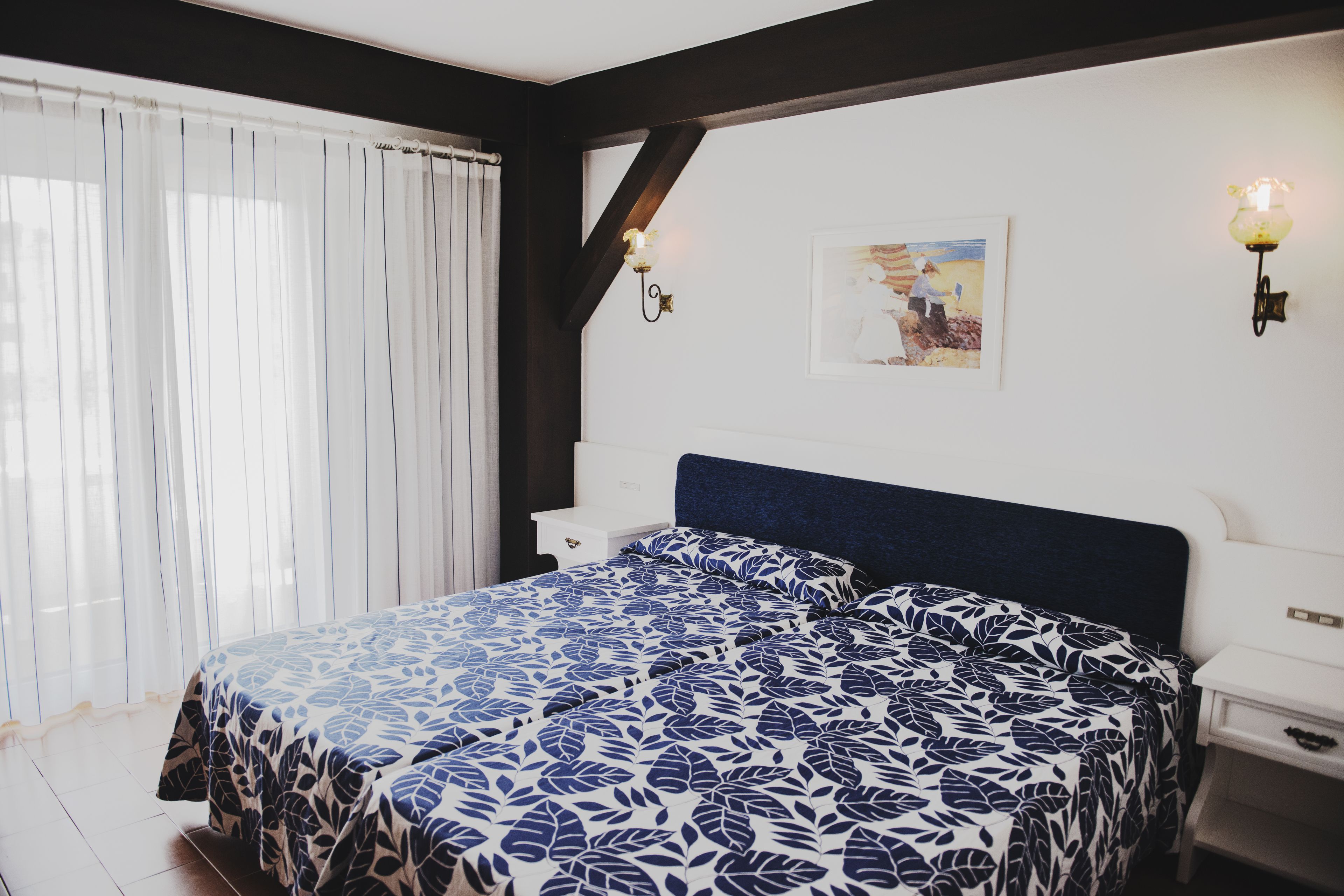 A bedroom with a large bed, white nightstands, a dark headboard, and a window with sheer curtains.