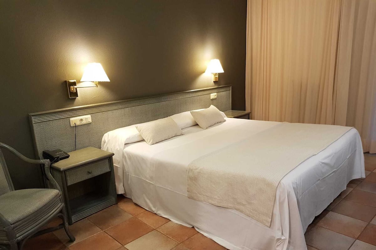 A neatly made bed with white linens, a nightstand with a lamp, and a chair.