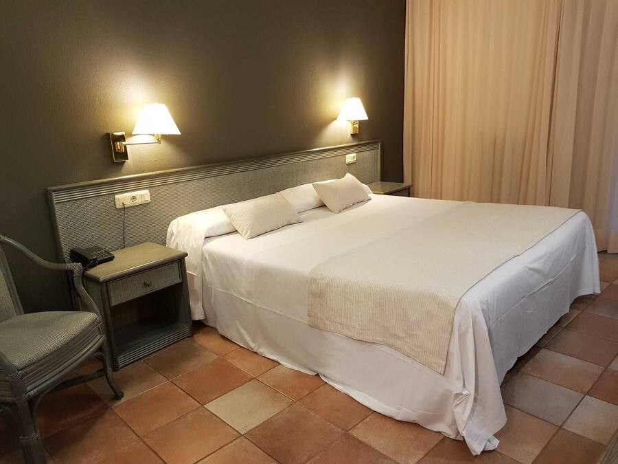 Een net opgemaakt bed met witte lakens, een nachtkastje met een lamp en een stoel.