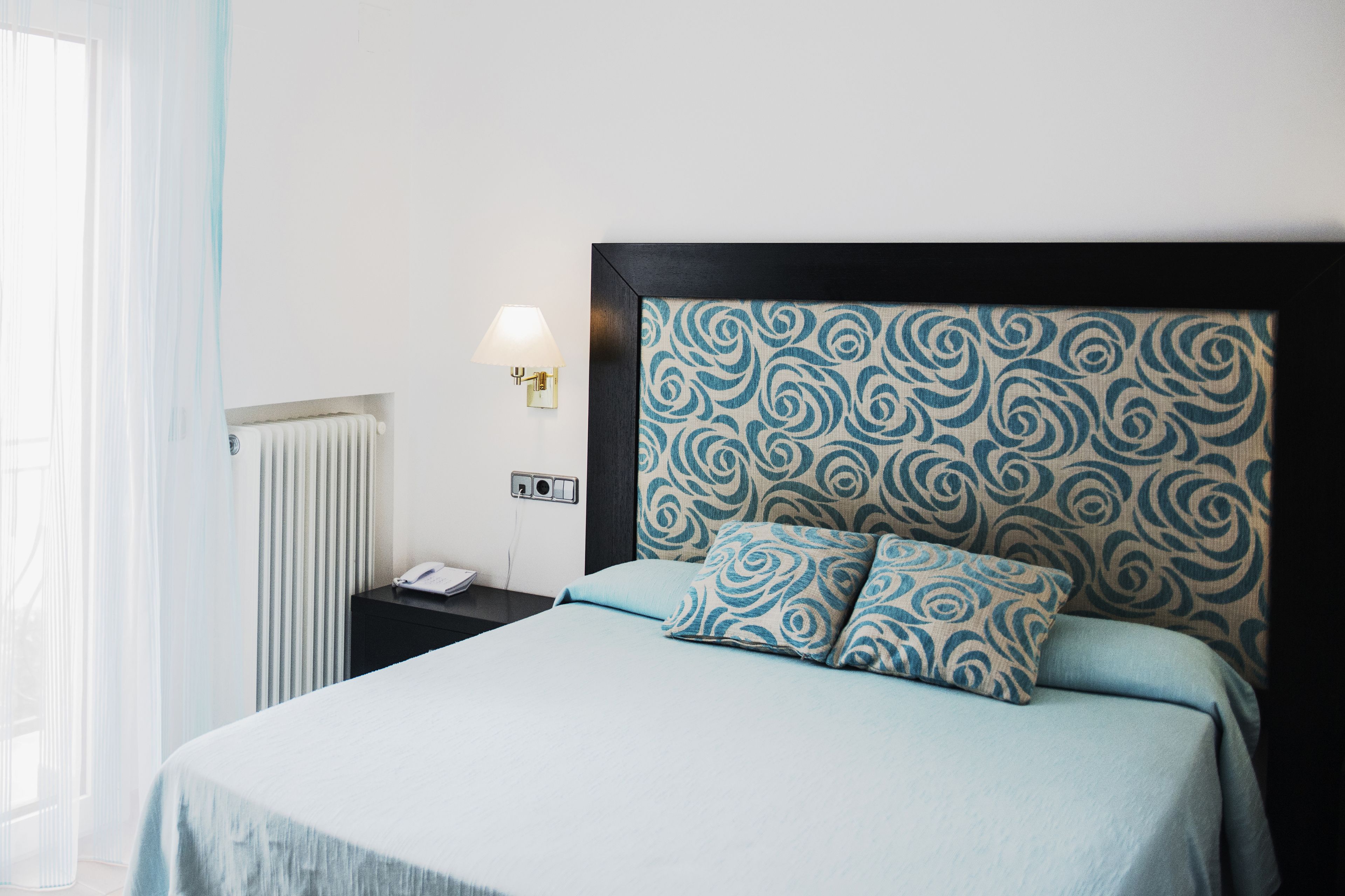 Ein ordentlich bezogenes Bett mit einem gemusterten Kopfteil, ein Nachttisch mit Lampe und ein Heizkörper an der Wand.