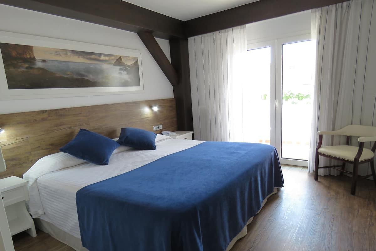 A hotel room with a large bed, a wooden headboard, a painting on the wall, a chair, and a view through the window.
