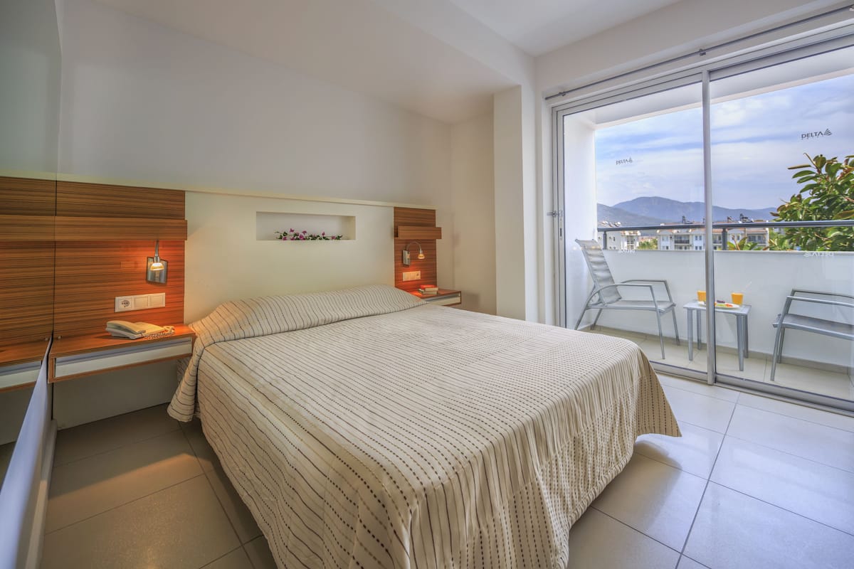 Een hotelkamer met een bed, nachtkastjes, een klein plankje met decoratieve items, en een glazen schuifdeur naar een balkon met stoelen en een tafel.