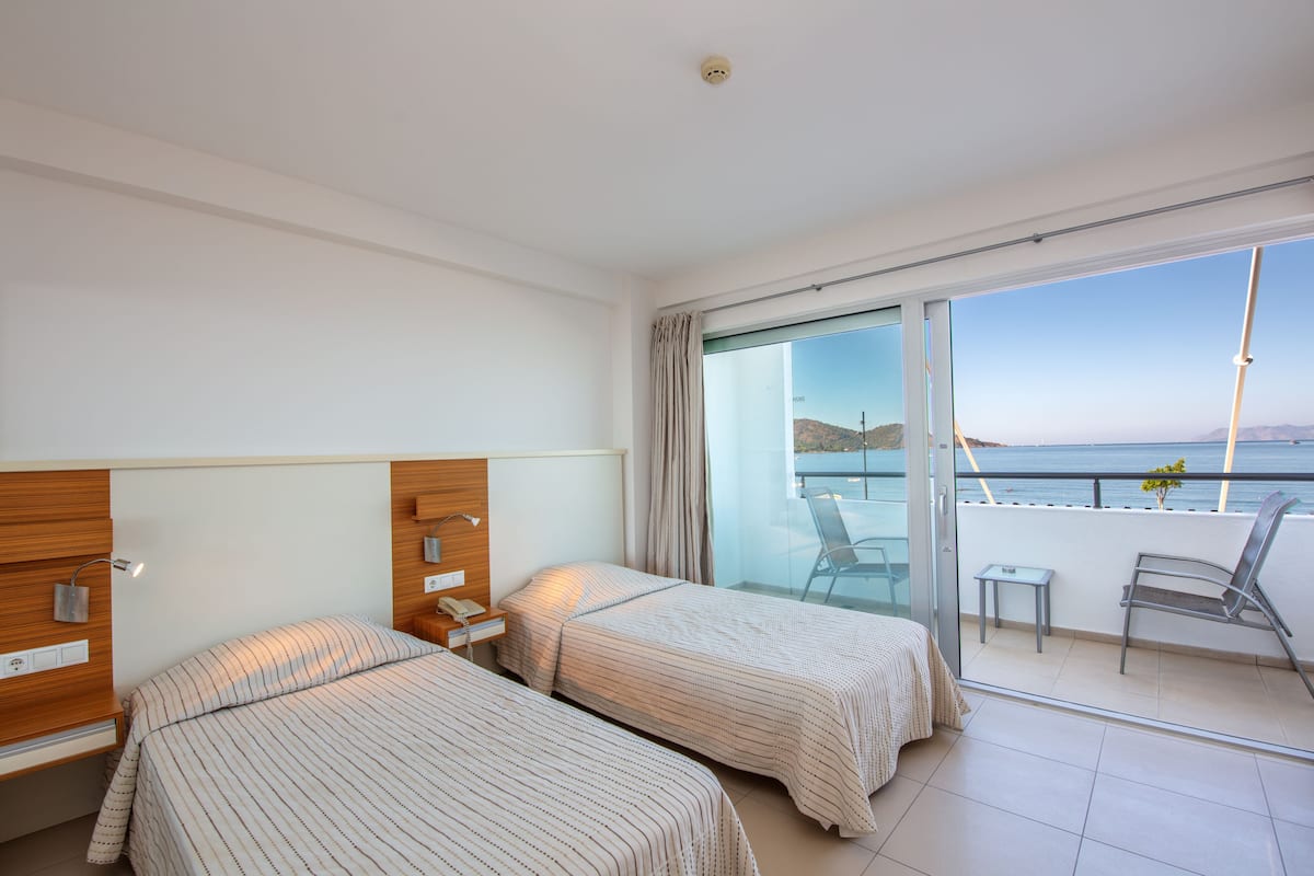 Een hotelkamer met twee bedden, een balkon met uitzicht op zee, en een klein tafeltje met stoel.