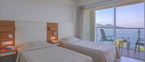 Habitación de hotel con dos camas, cabecero de madera, mesita de noche, una pequeña mesa con una bebida y un balcón con vista al mar.