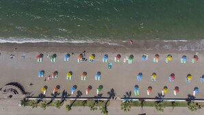 Vista aérea de uma praia com fileiras de guarda-sóis coloridos e cadeiras de praia, uma área de palco e palmeiras.