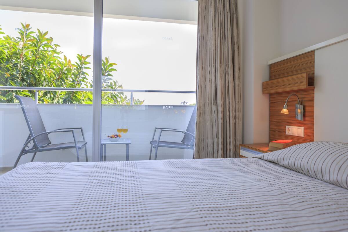 Hotelkamer met een bed, een tafeltje met een drankje, twee stoelen, een balkon met planten en een nachtkastje met een lamp.