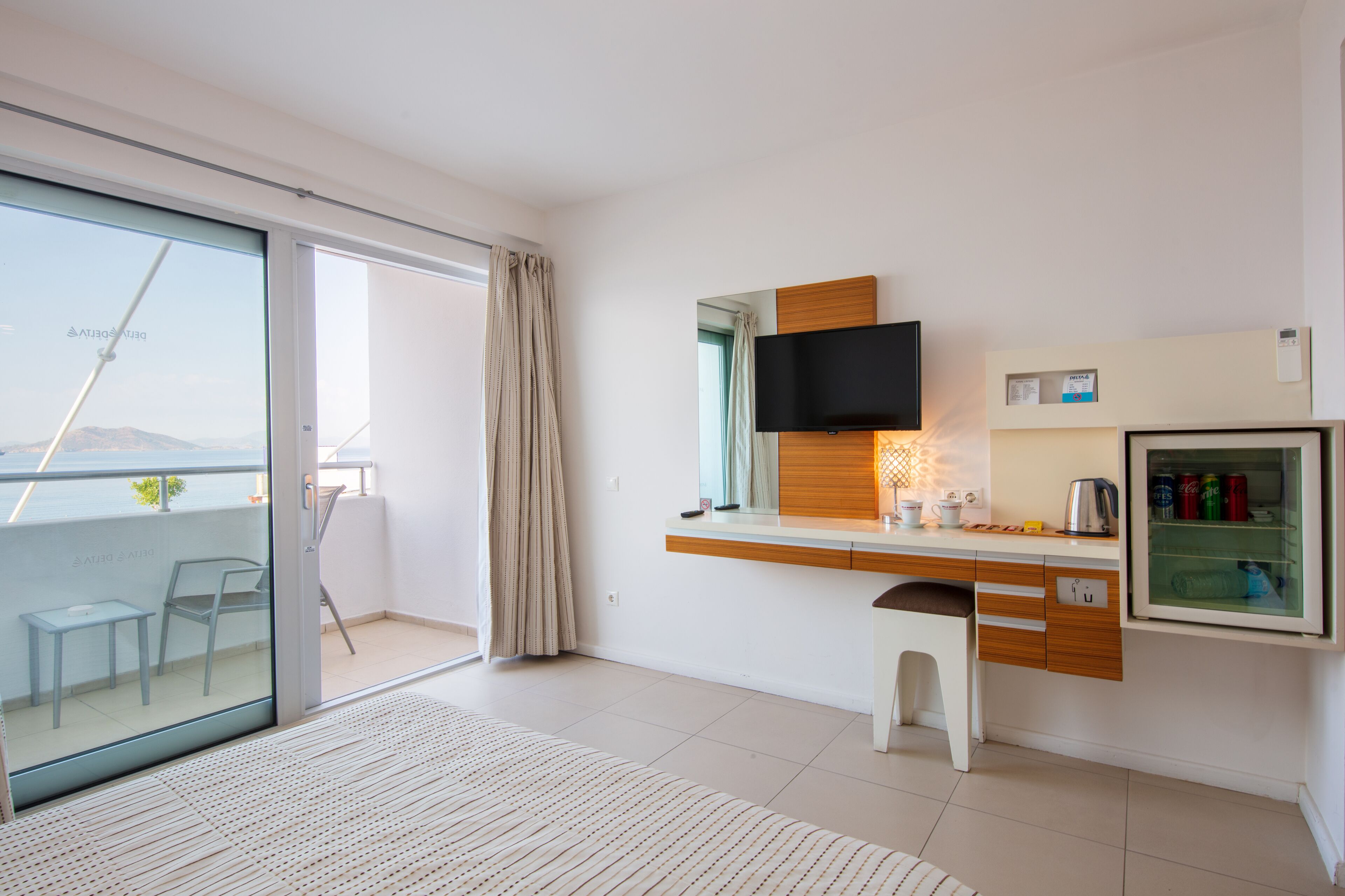 A modern hotel room with a sliding glass door to a balcony, a small desk with a flat-screen TV, a mini-fridge, and a glass cabinet with beverages.