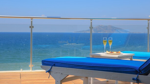 Uma varanda com duas cadeiras de descanso, uma mesa com um prato de comida e dois copos de vinho, e vista para o oceano e um barco ao longe.