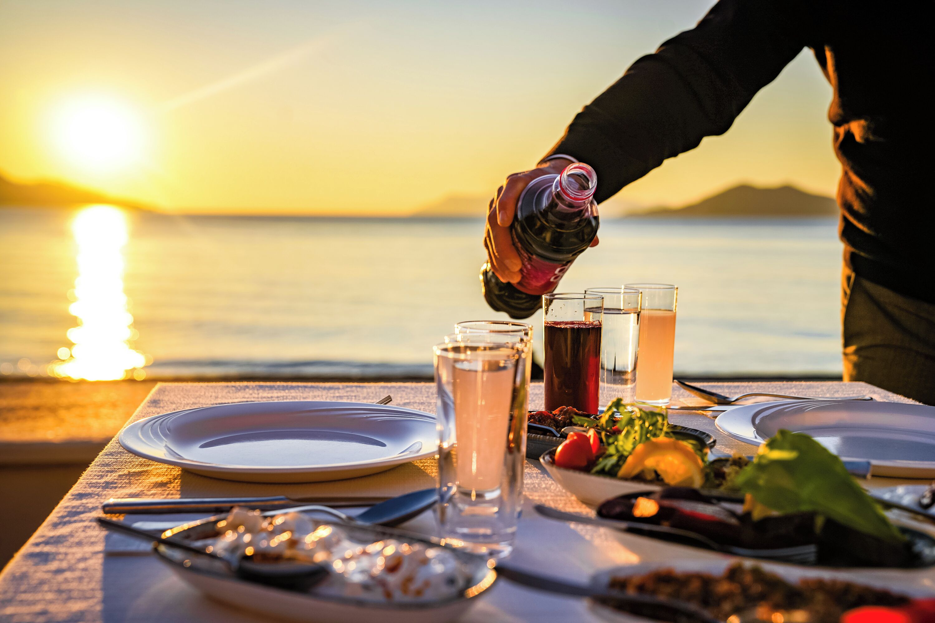 A table set for a meal with plates, cutlery, and glasses, overlooking a body of water at sunset.