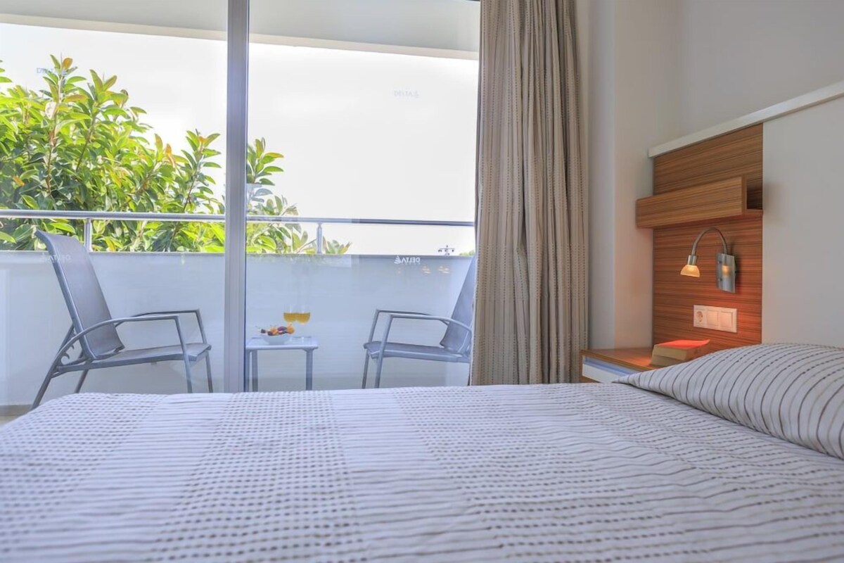 A hotel room with a bed, a small table with a drink, two chairs, a balcony with plants, and a wall-mounted light fixture.