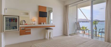 Habitación de hotel moderna con cama, televisor, minibar, taburete pequeño y balcón con vista al mar.