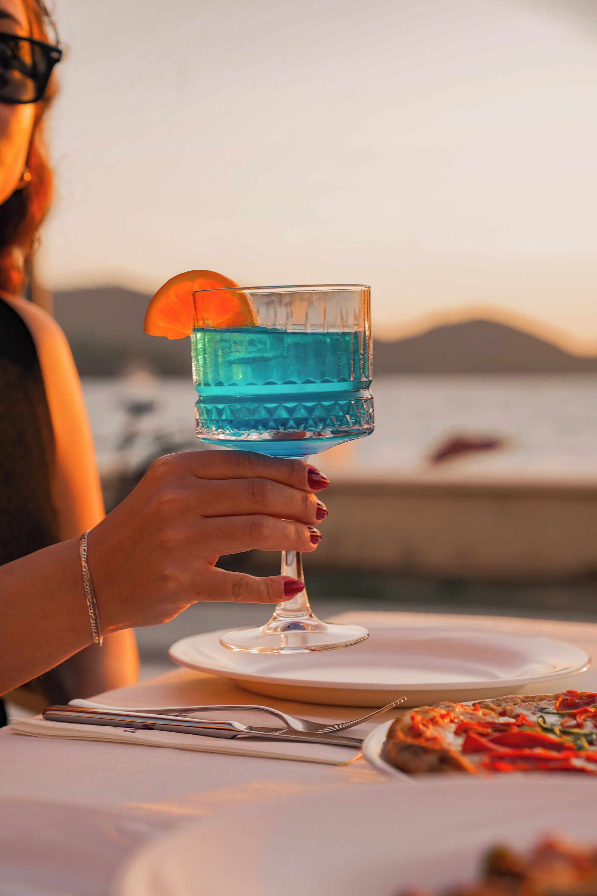 A person holding a blue cocktail glass with an orange garnish, a plate with a pizza, and a scenic view of the sea and mountains in the background.