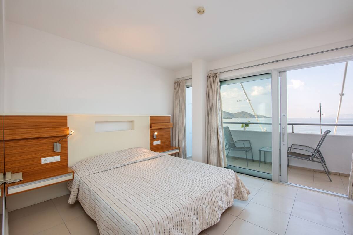 Een slaapkamer met een bed, een houten hoofdbord, nachtkastjes en een glazen schuifdeur die leidt naar een balkon met stoelen.