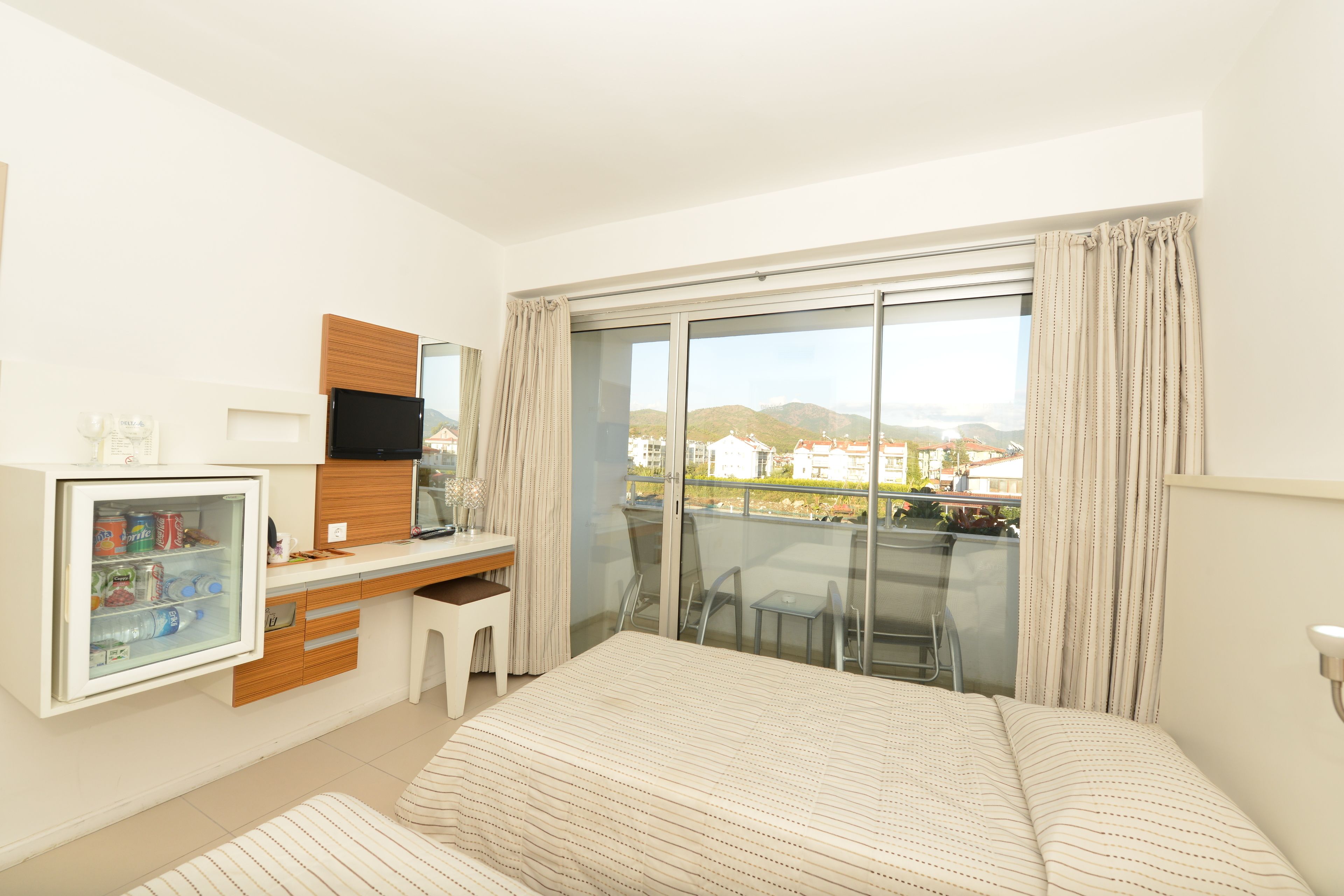 A hotel room with a bed, a desk with a computer, a mini-fridge, and a balcony with a view of the city.