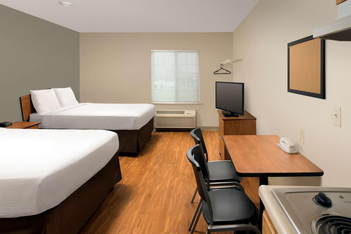 A hotel room with two beds, a desk with a chair, a television, a small table with a phone, and a kitchenette with a stove.