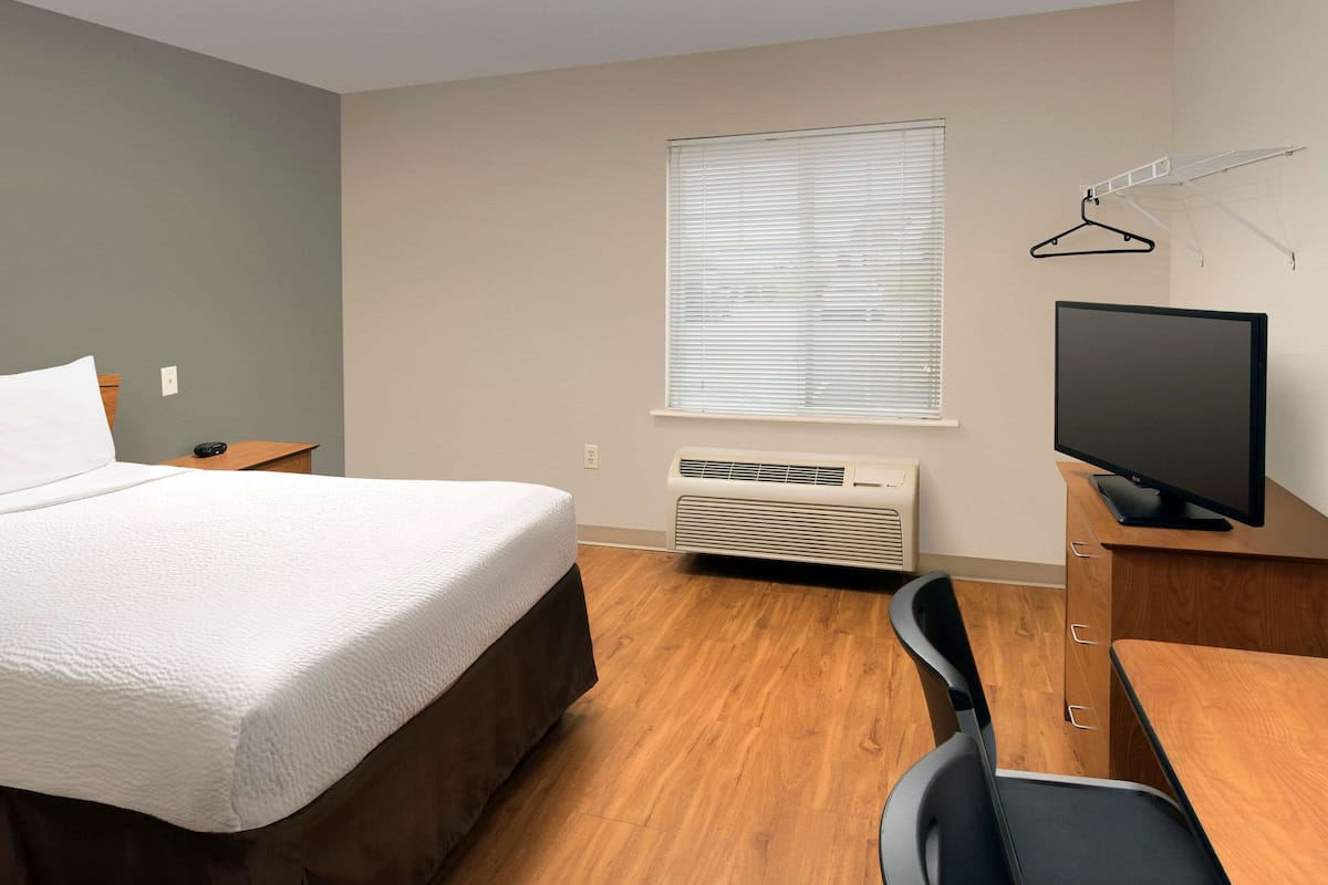 A hotel room with a bed, a desk, a chair, a television, a window with blinds, and an air conditioning unit.
