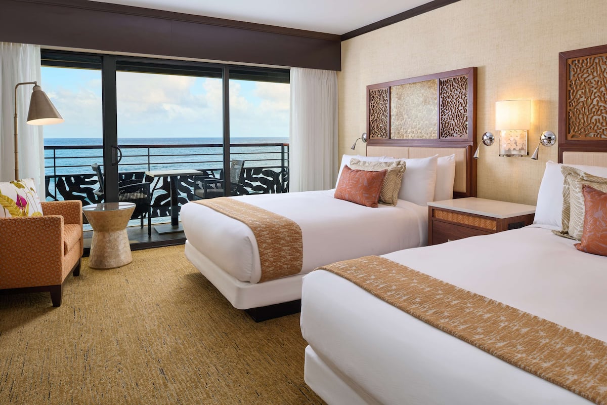 A hotel room with two beds, a sofa, a small table, and a balcony with a view of the sea.