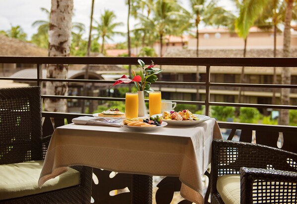 A dining table set with breakfast items, a vase with flowers, and two glasses of orange juice on a balcony with a view of palm trees and buildings. - Ko'a Kea Resort on Po'ipu Beach (Koloa)