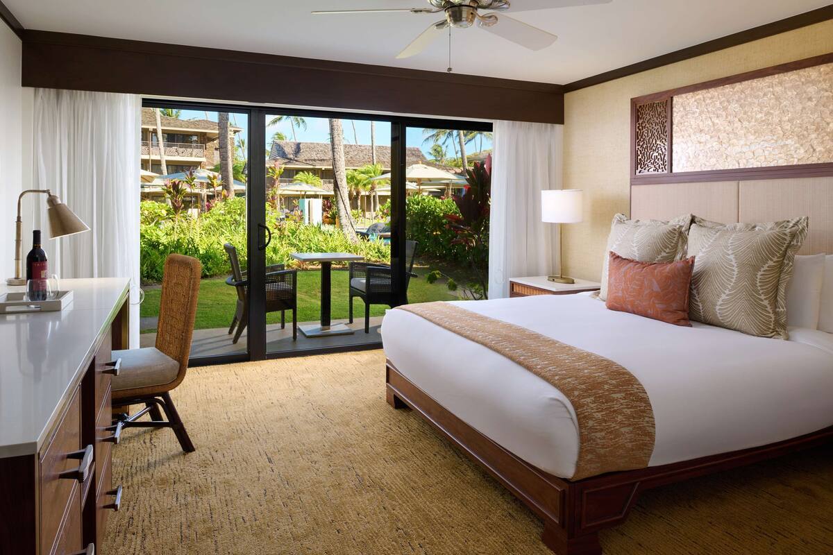 A hotel room with a large bed, a desk, a chair, and a view of a resort with palm trees and a pool.