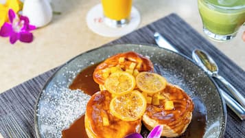 A breakfast plate with pancakes, syrup, and lemon slices. There is a glass of orange juice and a green drink on the table.