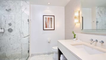 Un baño moderno con ducha de vidrio, suelo de mármol, lavamanos blanco y una obra de arte enmarcada en la pared.