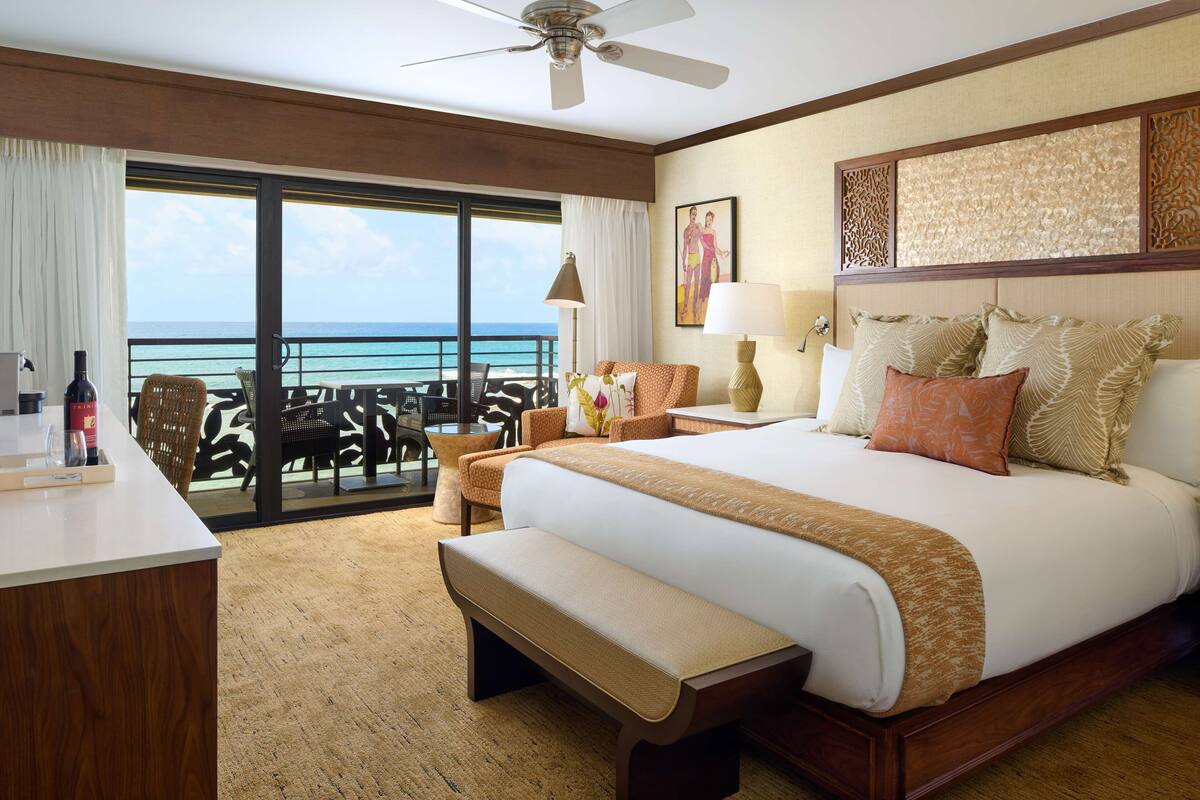 A hotel room with a large bed, a desk, a bench, a chair, a table, a lamp, and a view of the ocean.