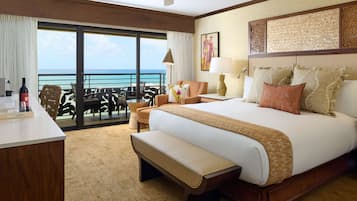 A hotel room with a large bed, a desk, a bench, a chair, a table, a lamp, and a view of the ocean.