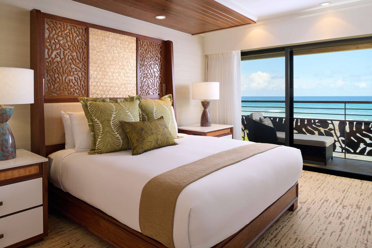 A hotel room with a large bed, a nightstand with a lamp, a view of the ocean through a balcony, and a decorative wall panel.
