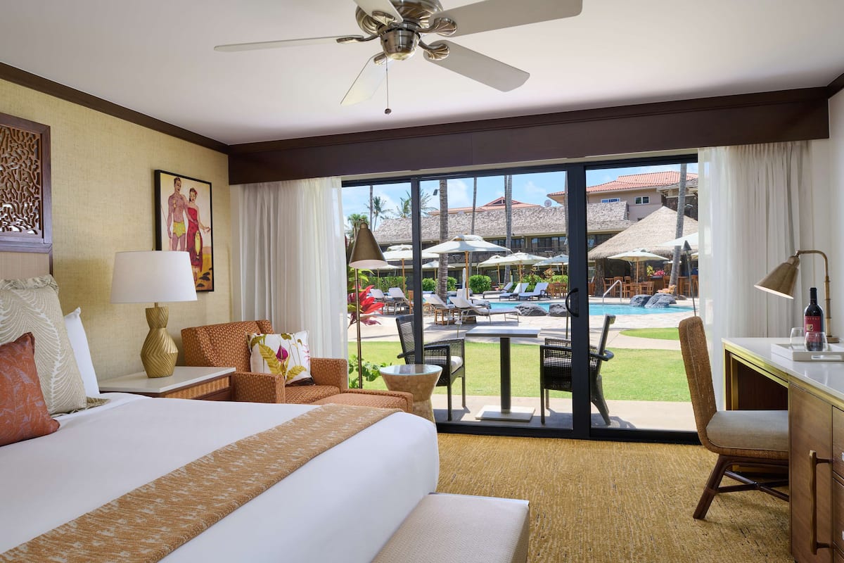 A hotel room with a large bed, a desk, a chair, and a view of a pool and outdoor seating area.