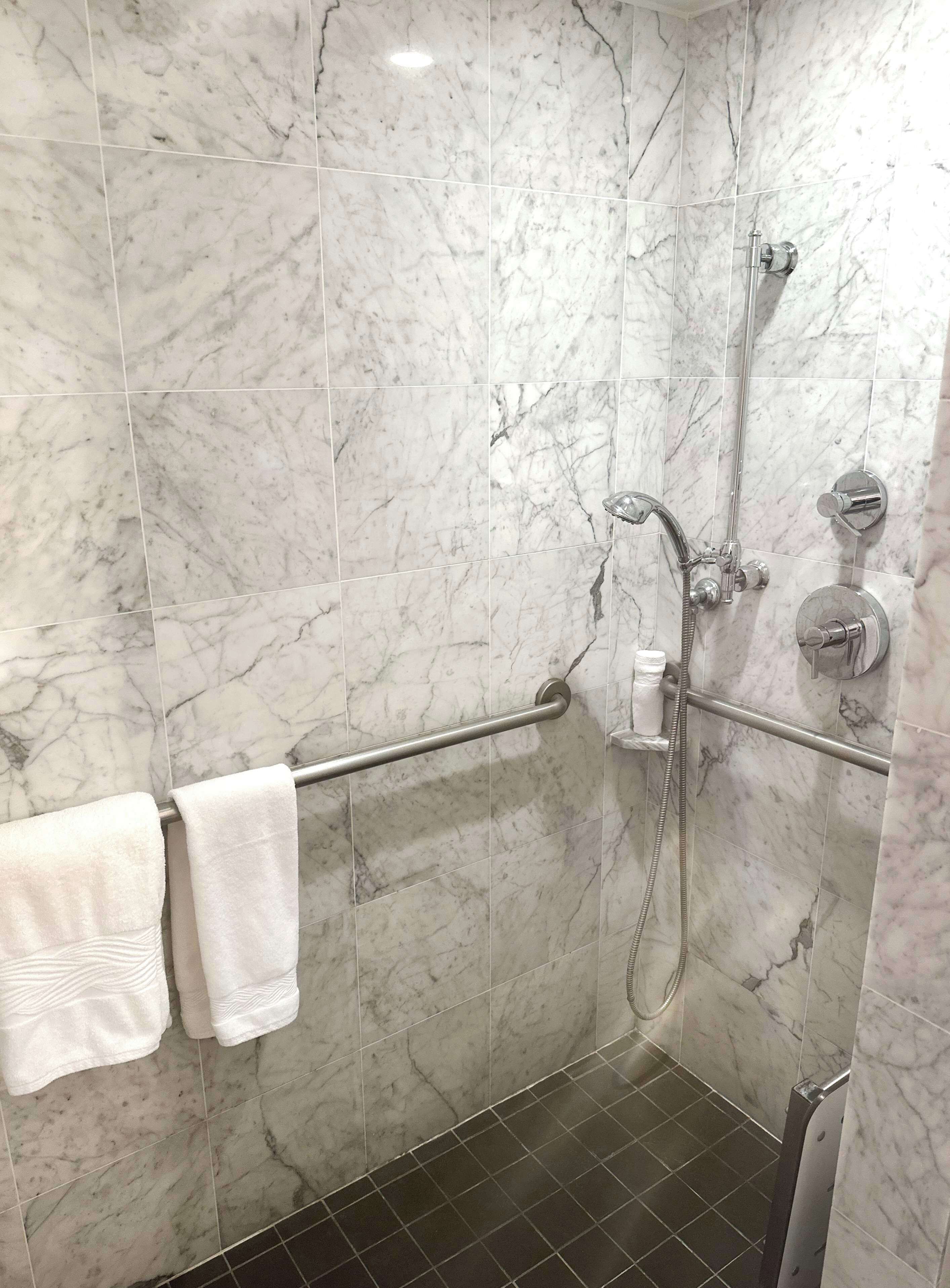 Une salle de bain aux murs recouverts de marbre, une douche avec un pommeau de douche amovible, une barre d’appui et deux serviettes sur un porte-serviettes.