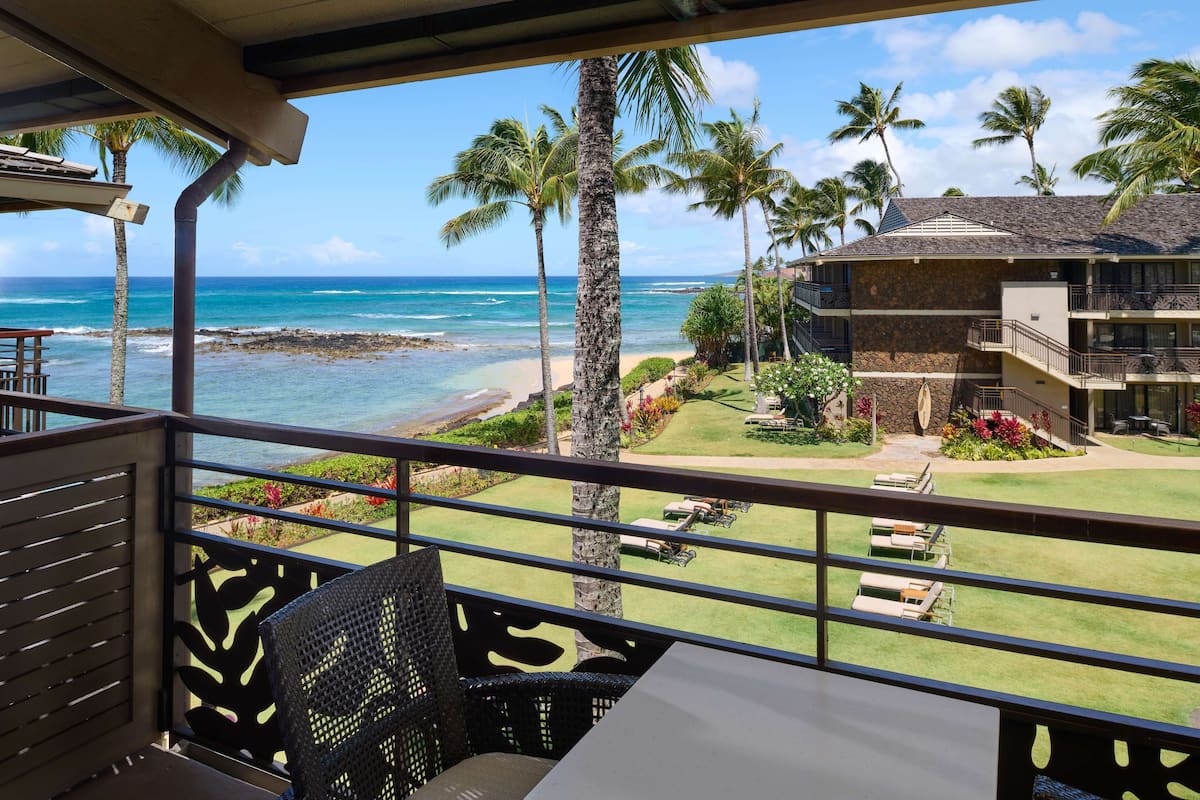 A balcony with a view of the ocean, palm trees, and a building with a stone facade.