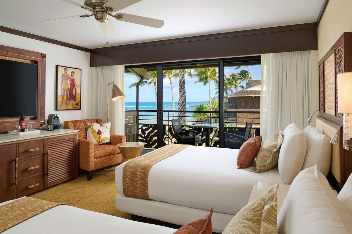 A hotel room with a large bed, a desk with a chair, a sofa, a TV, and a balcony with a view of the ocean.