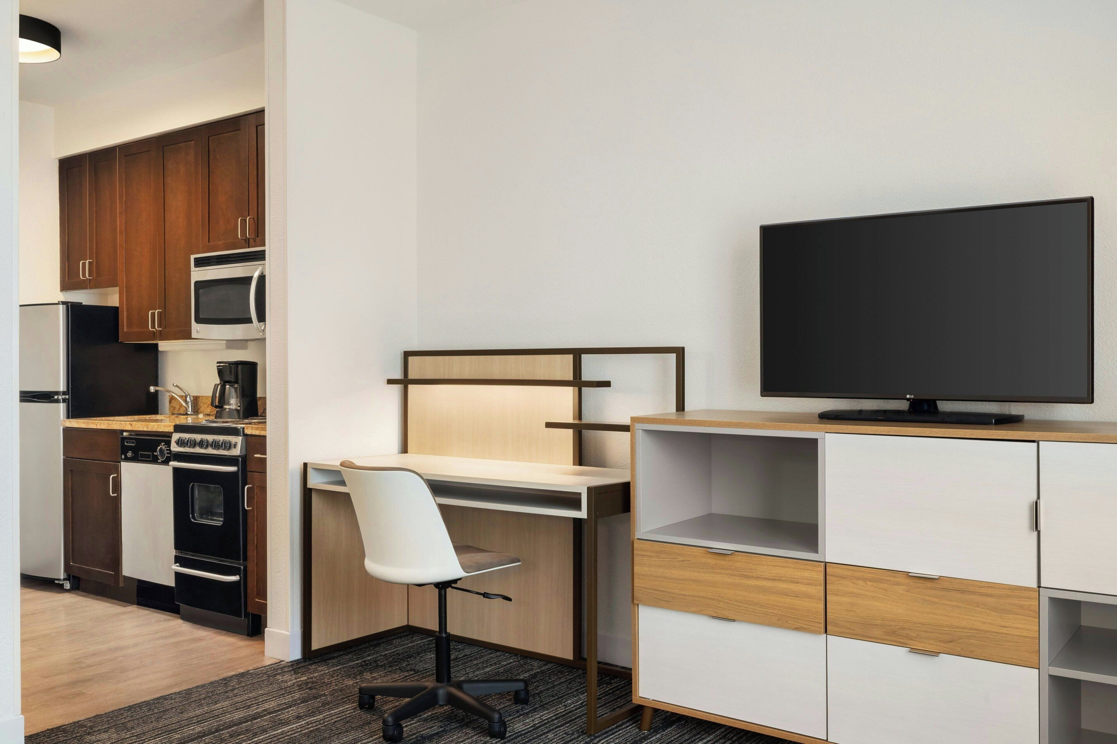 A modern kitchen with a microwave, stove, and coffee maker. There is a desk with a chair and a television on a stand.