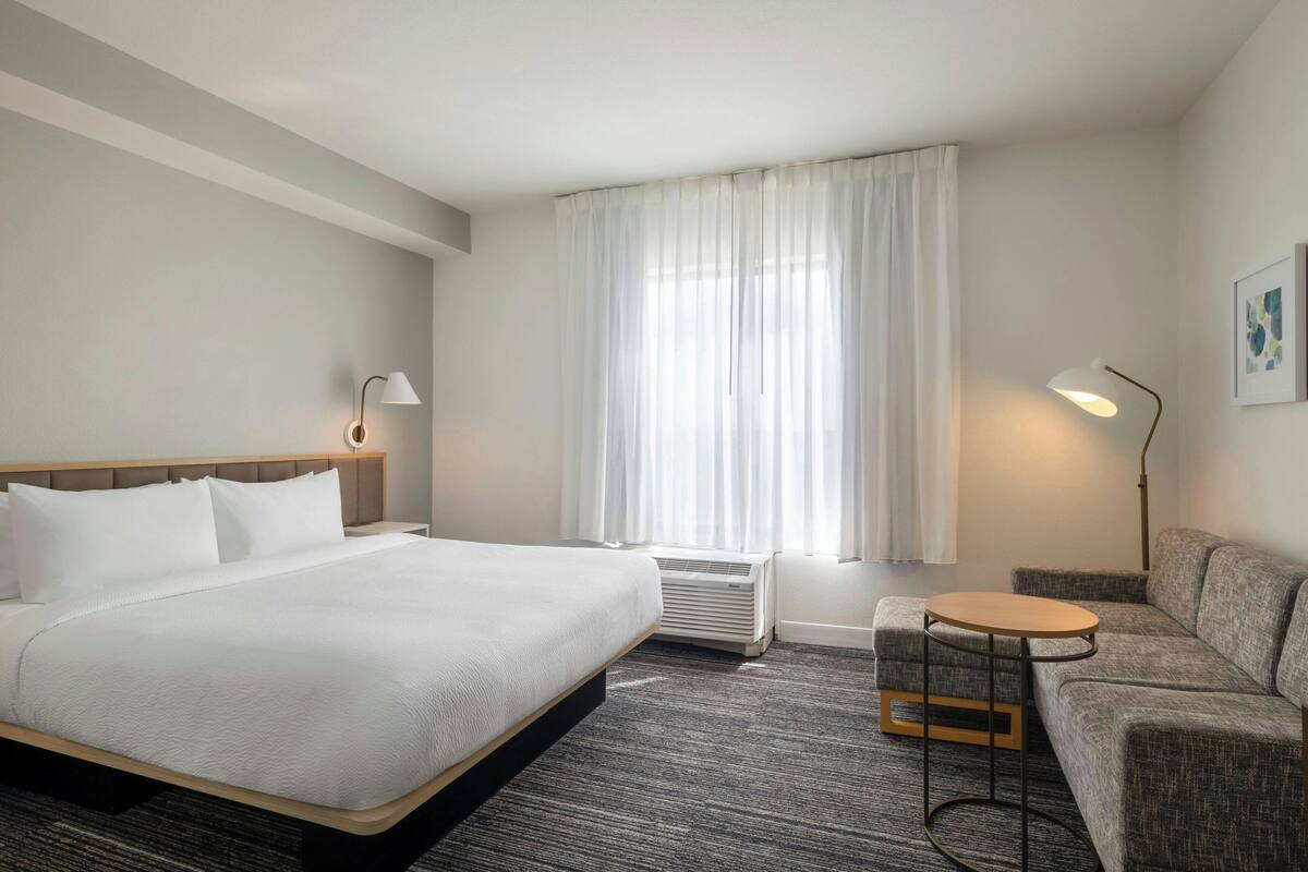 A hotel room with a bed, a sofa, a round table, a standing lamp, and a window with curtains.