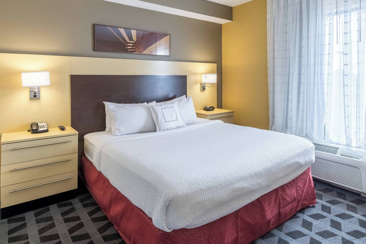 A hotel room with a large bed, two bedside tables with lamps, a headboard, a painting above the bed, and a window with curtains.