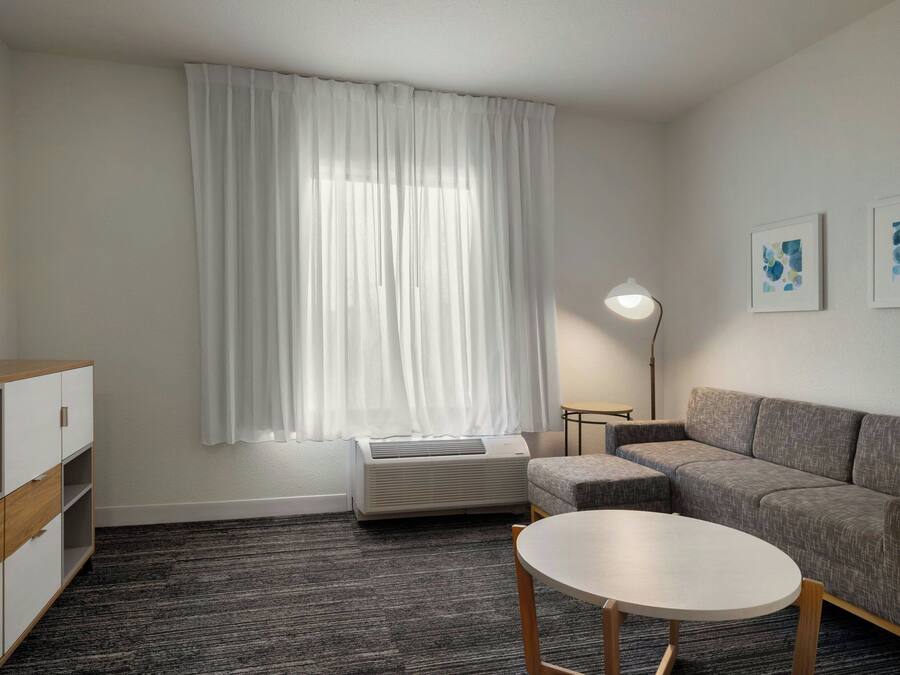 Et moderne værelse med sofa, et lille bord, fjernsyn på et skab, aircondition og et vindue med gardiner.