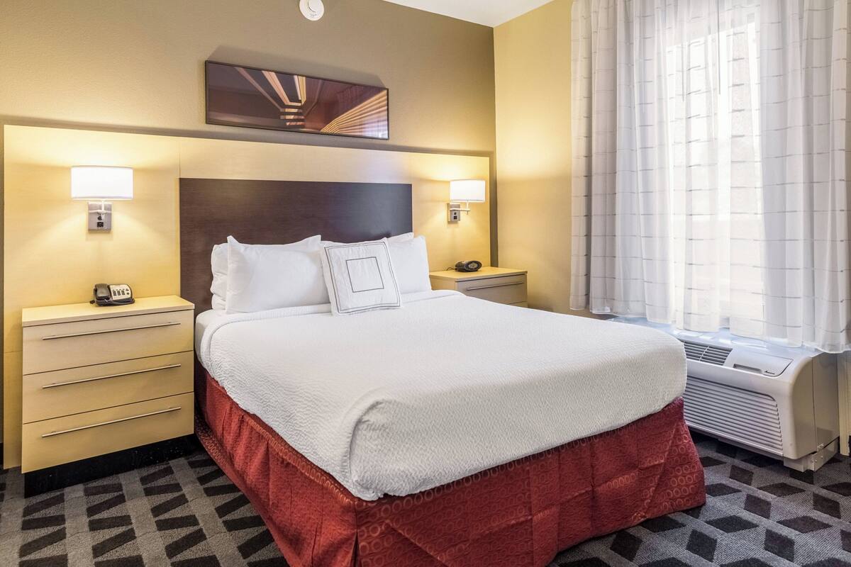 A hotel room with a large bed, two bedside tables, a wall-mounted artwork, and a window with curtains.