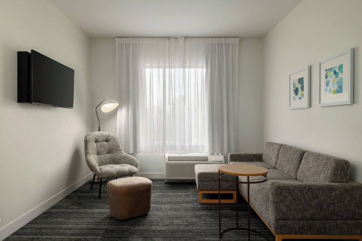 A modern hotel room with a grey sofa, a smaller armchair, a round table, a flat-screen TV, and a large window with sheer curtains.