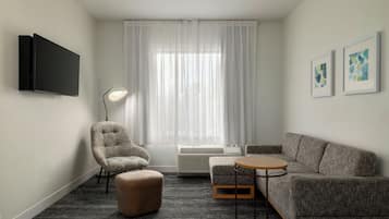 A modern hotel room with a grey sofa, a smaller armchair, a round table, a flat-screen TV, and a large window with sheer curtains.