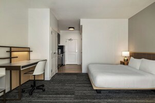 Quarto de hotel com cama, escrivaninha, cadeira e uma cozinha compacta ao fundo.
