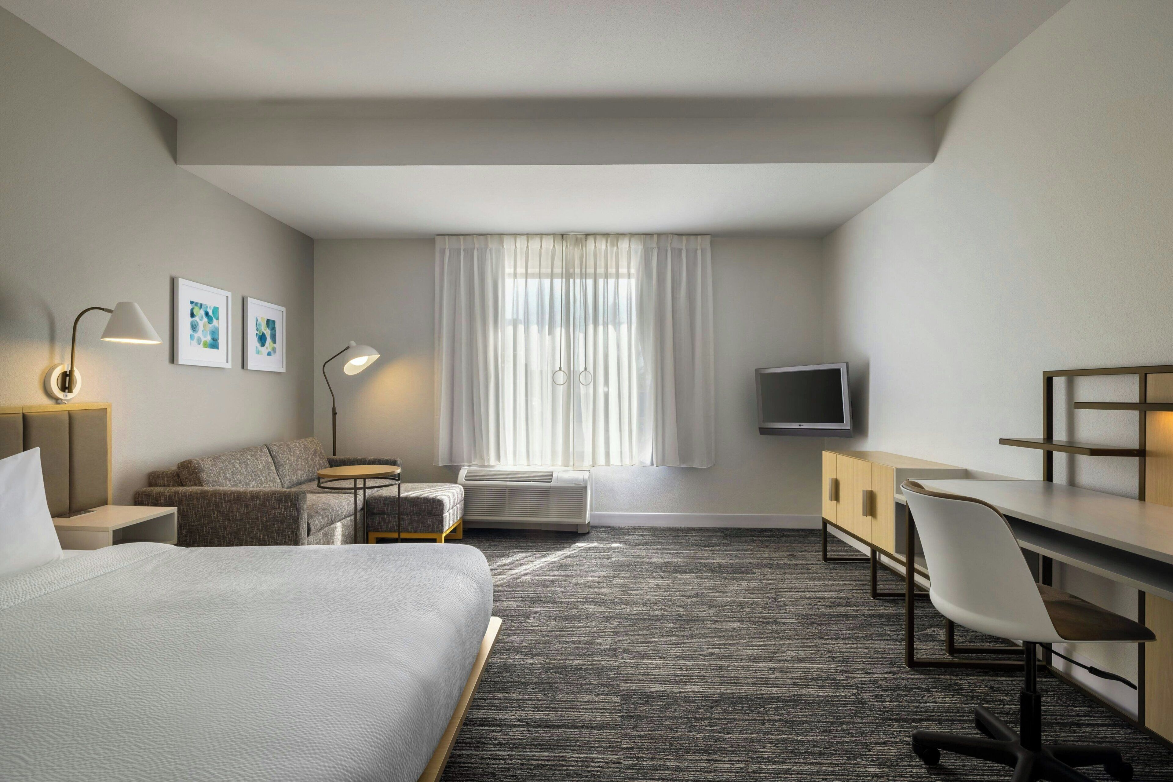 Une chambre d’hôtel avec un lit, un bureau, une chaise, une télévision et une fenêtre avec des rideaux.