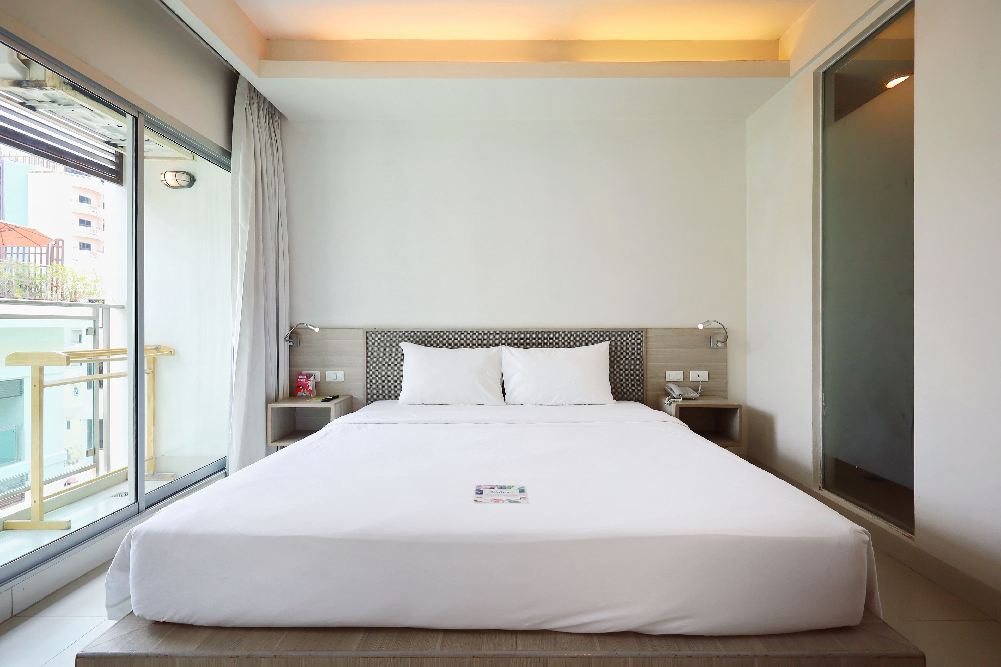 A hotel room with a large bed, a bedside table, a nightstand, a window with curtains, and a view of buildings outside.