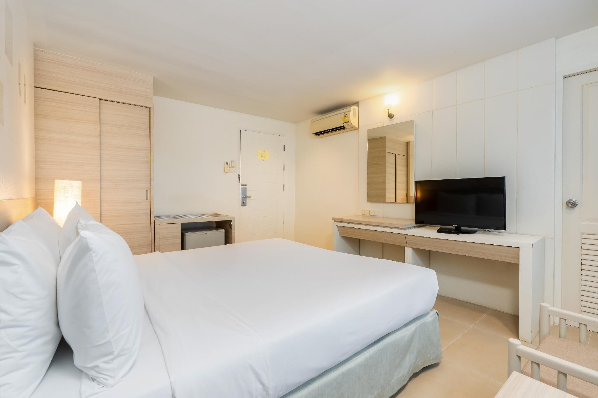 A hotel room with a bed, a desk with a computer, a TV, and an air conditioning unit.