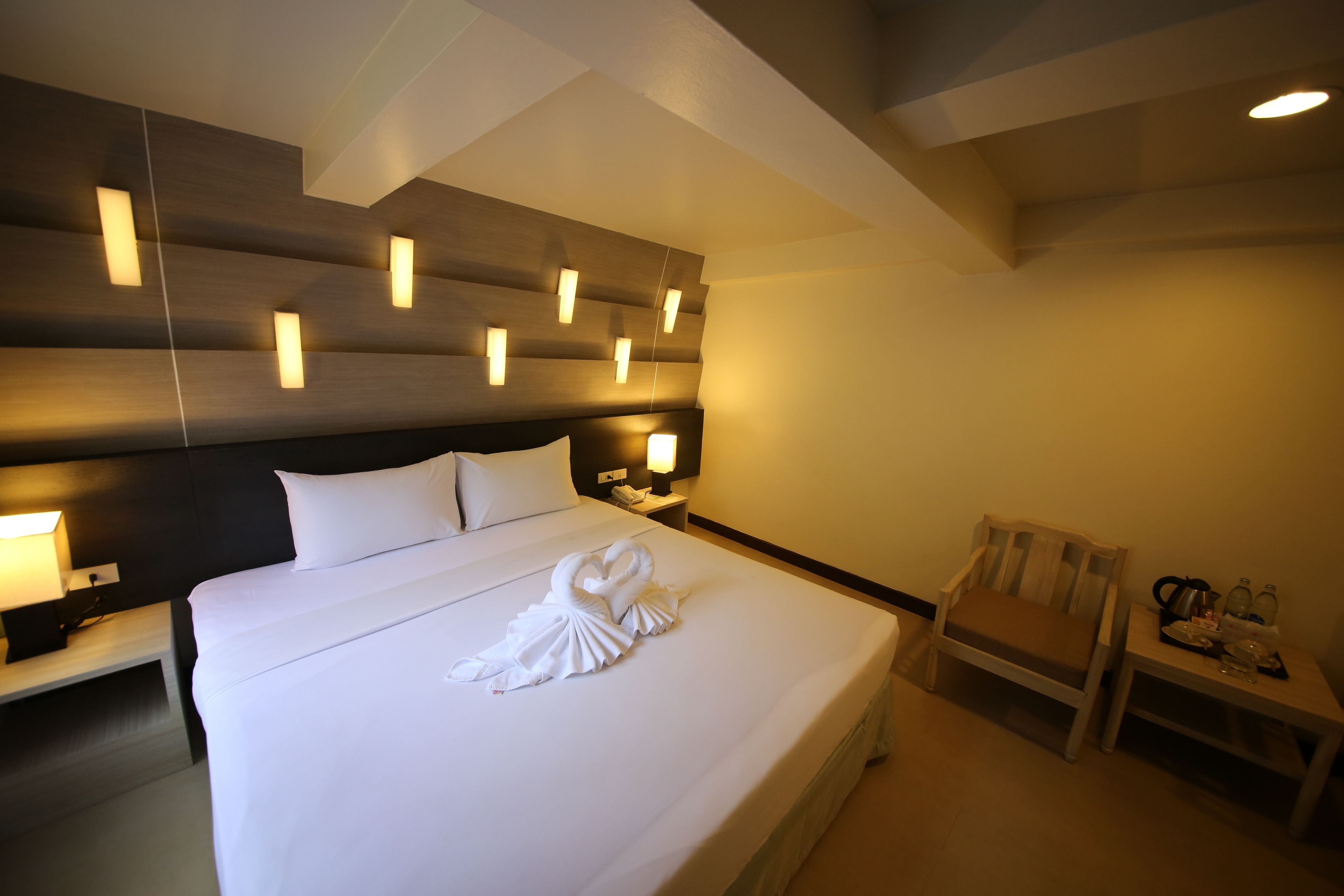 A hotel room with a large bed, a wooden headboard, bedside tables with lamps, a chair, and a small table with a kettle and cups.