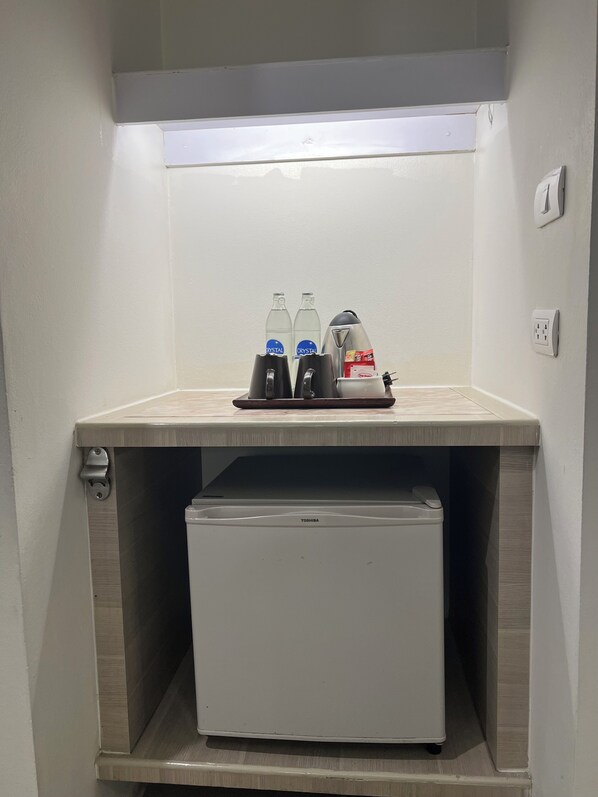A corner with a white refrigerator, a small shelf with two bottles labeled 'CRYSTAL,' a kettle, and a power outlet. - Sunshine Hotel And Residences (Pattaya)