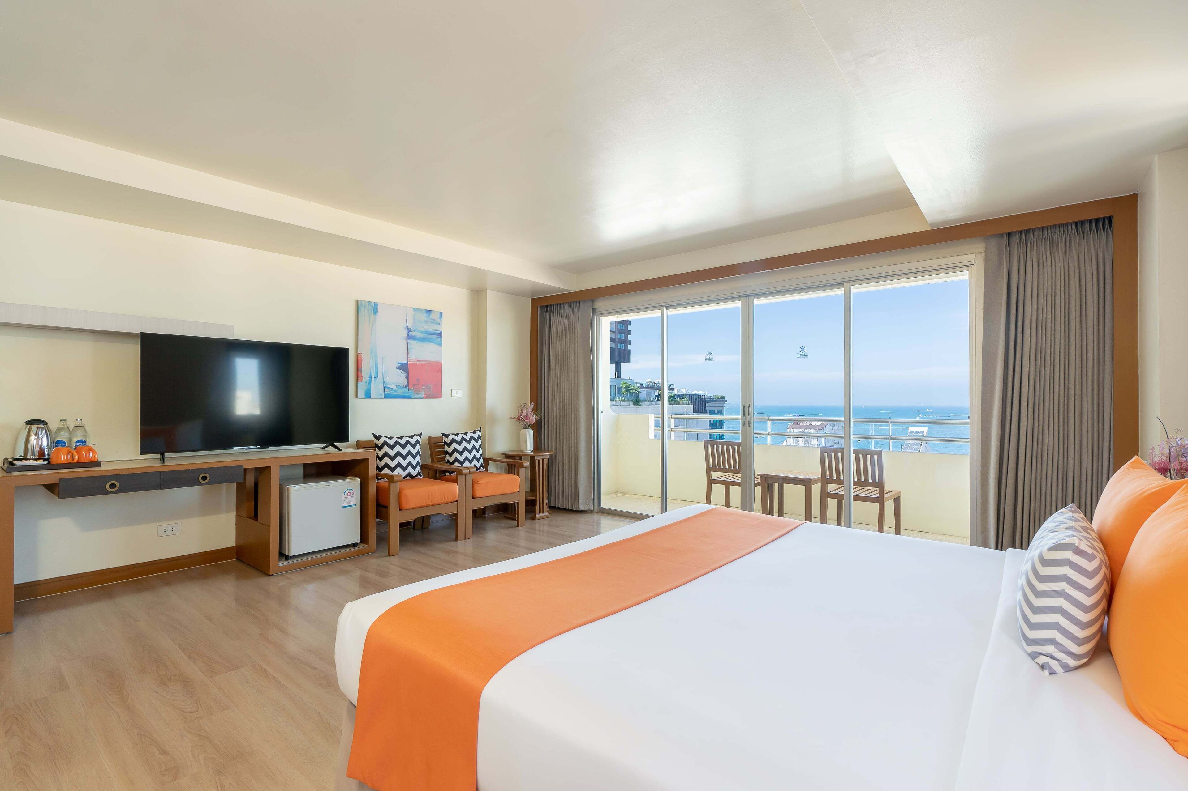 Ein Hotelzimmer mit einem großen Bett, einem Schreibtisch mit Fernseher, einer kleinen Kühlbox und einem Balkon mit Meerblick.
