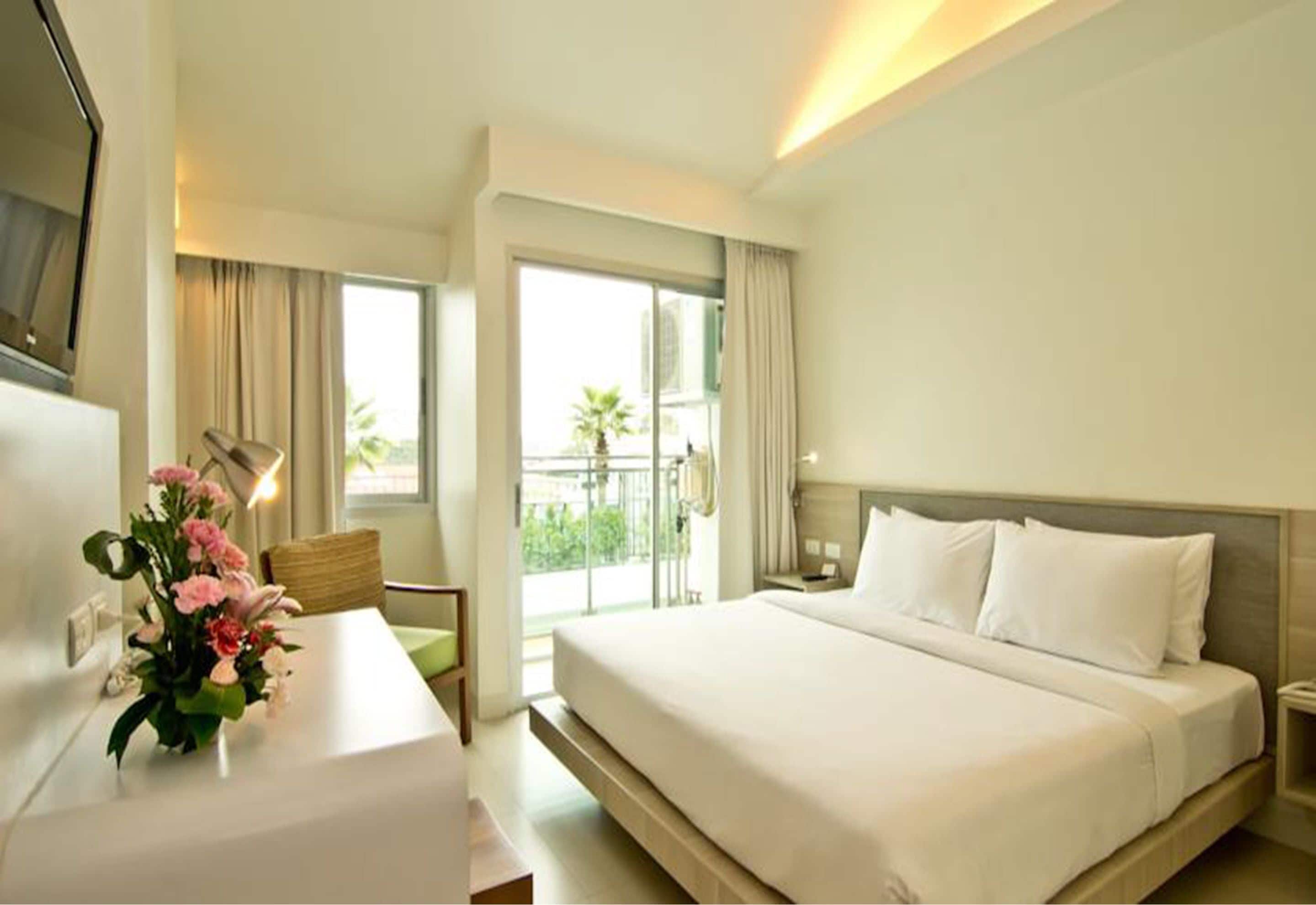 A hotel room with a bed, a desk with a chair, a TV, and a balcony with curtains.